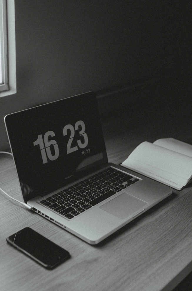 Grayscale Photo of Macbook on Wooden Table with Time Displayed on Screen